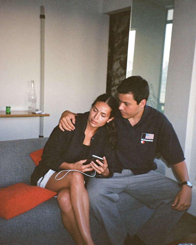 Pareja mexicana en sus treinta sentada en el sofá; ella le pregunta algo a su celular mientras él se acerca a escuchar la respuesta